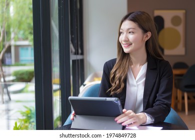 Asian Professional  Working Woman In A Black Suit Is Working On An Ipad Or Tablet On The Table Smiling Happily In The Office And Working At Home.