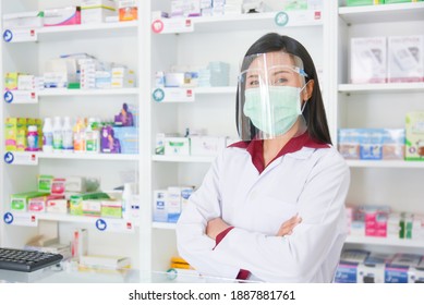 Asian professional young pharmacist wearing faceshield and mask while standing at drugstore.