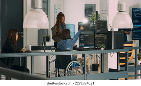 Asian programer with chronic impairment working on software code with it engineer in agency office. App developer with disability in wheelchair using html script on temrinal window.