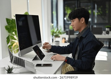 An Asian Programmer at his desk using multiple devices for software development in a contemporary tech office.