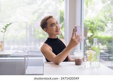 Asian queer LGBT community supporter man with mustache wearing lipstick and jumpsuit in a bright, modern cafe with iced coffee and greenery using mobile phone while taking selfie or video call - Powered by Shutterstock