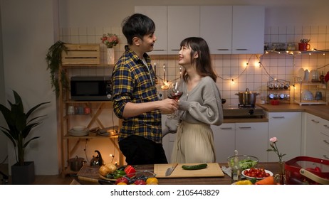 Asian Romantic Couple Flirting With Each Other With Wine On Valentine’s Night Date At Home In The Kitchen. The Man Gently Touching Girlfriend’s Shoulder While Talking With Hand Gestures