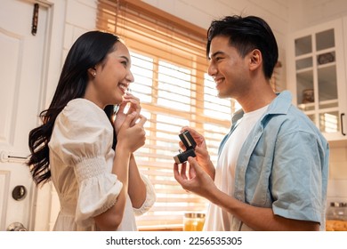 Asian romantic man making surprise proposal of marriage to girlfriend. Attractive young male and proposing to beautiful happy woman, with wedding ring enjoying surprise engagement in kitchen at home.