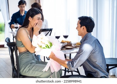 Asian romantic man making surprise proposal of marriage to girlfriend. Attractive young male and proposing to beautiful happy woman, with flower, enjoying surprise engagement in cafe restaurant. - Powered by Shutterstock