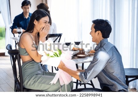 Asian romantic man making surprise proposal of marriage to girlfriend. Attractive young male and proposing to beautiful happy woman, with flower, enjoying surprise engagement in cafe restaurant.