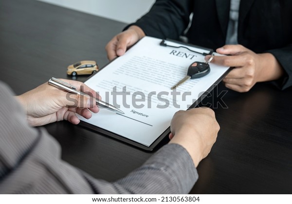 Asian\
salesman holding rental contract and hand over car key to client\
while he is holding pen to signing on the contract car trade or car\
loan or car rental on the table in the\
office