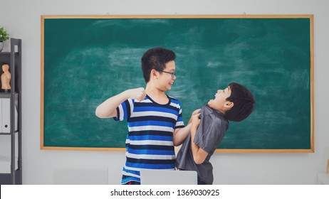 Asian School Boy Quarreling, Punching, Bullying, Weak Friend On Blackboard In Classroom Background With Angry Face.Bully And Physical Abuse Of Children And Kid Student Concept.