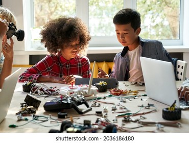 Asian school kid boy helping African child girl student using tablet computer studying programming robots learning making robotic cars sitting at table at STEM education coding engineering class.