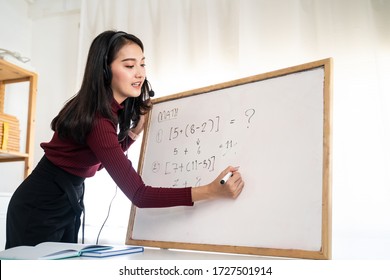 Asian school young woman teacher working from home teach online math subject to student studying from home. Girl writes on whiteboard, talk on headphone. Remote education class during covid19 pandemic