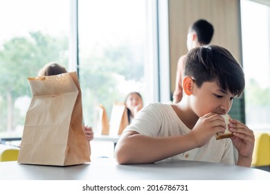 Asian Schoolboy Eating Sandwich Alone In Dining Room Near Blurred Teenagers