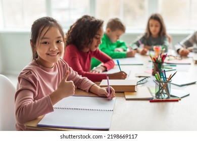 Asian Schoolgirl Gesturing Thumbs Up Smiling To Camera Taking Notes Learning With Multicultural Kids In Modern Classroom At School. Education Concept. Selective Focus - Powered by Shutterstock