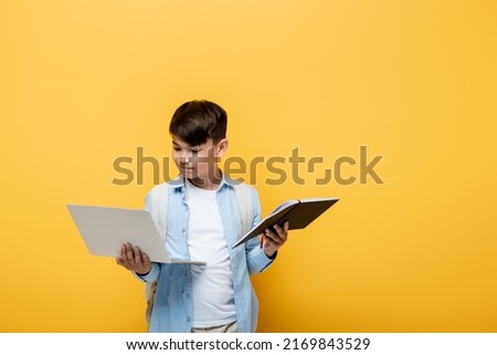 Asian schoolkid holding laptop and notebook isolated on yellow