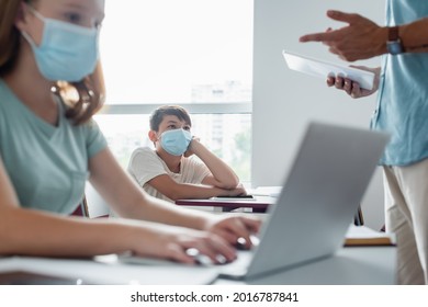 Asian Schoolkids In Medical Mask Looking At Blurred Teacher With Digital Tablet