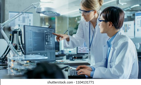 Asian Scientist Sitting at His Desk Consults Senior Engineer about Sophisticated Coding and Programming. In the Background Computer Science Research Laboratory with Robotic Arm Model.
