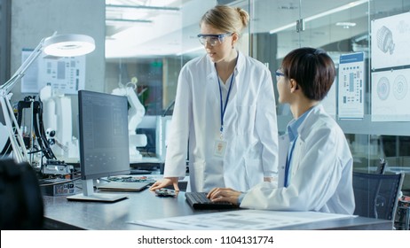 Asian Scientist Sitting At His Desk Consults Senior Engineer About Sophisticated Coding And Programming. In The Background Computer Science Research Laboratory With Robotic Arm Model.