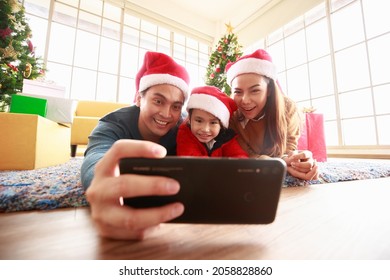 Asian Selfie Holding Smartphone Taking Picture. Happy Young Family With Kids Fun Celebrating Christmas. Christmas Time. My Dad, Mom, And Daughter In Santa Hats Lie About In Front Gift Box At Home