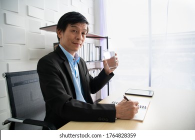 Asian Senior Businessman Stay In His Office, Elder Employee Drinking A Water To Refresh Himself During Working. Health And Work Life Balance Concept In Senior People. Businessman Relaxing And Drinking