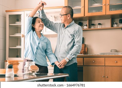 Asian  Senior Couple Is Dancing And Smiling While Cooking Together In Kitchen.