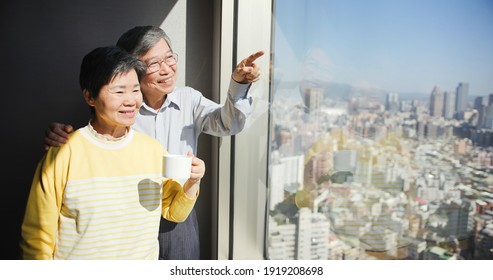 Asian Senior Couple Are Looking At City View And Chatting Near Window Happily While Traveling