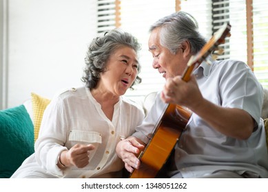 Asian senior Couple singing and playing acoustic guitar together. Elderly man and woman having fun while sitting on couch in the living room at their house. Retirement, life, Lifestyle, Carefree.