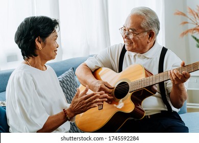 Asian senior couple sitting on the sofa and playing acoustic guitar together. Happy smiling elderly woman clapping hands while old 70s guitarist husband singing. Enjoying retirement life at home