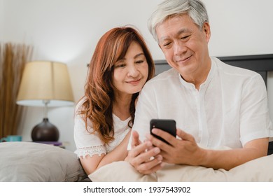 Asian senior couple using mobile phone at home. Asian Senior Chinese grandparents, husband and wife happy after wake up, watching movie lying on bed in bedroom at home in the morning concept.
