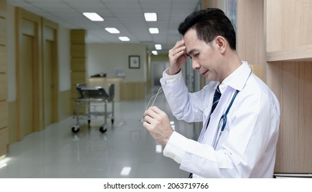 Asian Senior Doctor Man Wearing Medical Uniform Standing Against Bookshelf In Hospital Corridor While Take Off Glasses With Tired Rubbing Nose And Eyes Feeling Fatigue And Headache