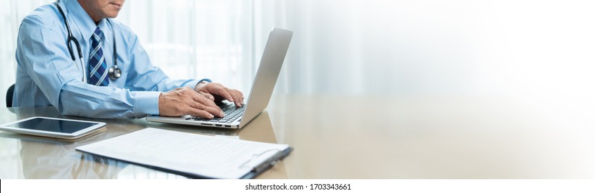 Asian Senior Doctor Using Laptop Computer, Digital Tablet, Clipboard In Medical Room. Elderly Physician In Blue Shirt Sitting Working At Workplace In Hospital. Banner, Web, Panoramic, Copy Space.