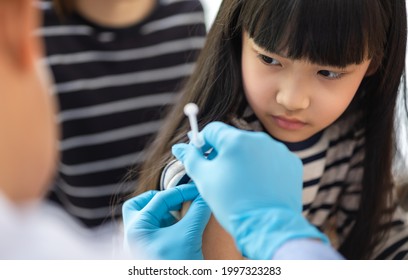 Asian  Senior Doctor Wearing Gloves And Isolation Mask Is Making A COVID-19 Vaccination In The Shoulder Of Child Patient With Her Mother At Hospital.closed Up Photo.