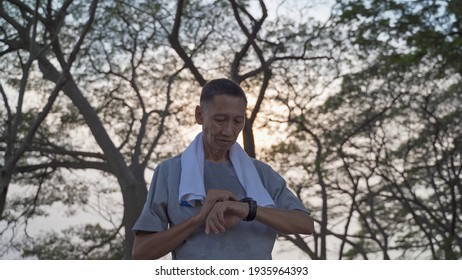 Asian Senior Elderly Man Touching Smart Watch Activity Tracker At Park Under Tree. Finger Tapping Sport Watch For Calories And Heart Rate Information. Slow Motion Orbit Shot. Health Care Concept.