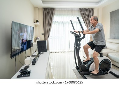 Asian Senior Elderly Man Workout Cardio On Cycling Exercise Bike Machine At Home For Well Being. Active Older Male Doing Lockdown Activity Follow Instruction Video From Online Fitness Trainer In House