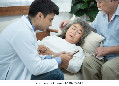 Asian Senior Elderly Old Patient Woman Laying Down On Sofa In Home Waiting For Result From Smart Young Caucasian Doctor Smartphone While Check Up In Home With Grandfather Take Care Beside.