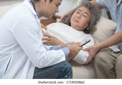 Asian Senior Elderly Old Patient Woman Laying Down On Sofa In Home Waiting For Result From Smart Young Caucasian Doctor Smartphone While Check Up In Home With Grandfather Take Care Beside.