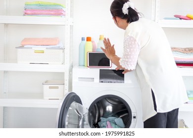 Asian Senior Elderly Old Woman Housewife Doing Laundry At Laundry Room During Looking At Tablet, Grandma Using Tablet In Learning New Things While Doing Housework And Cleaning Clothes At House.