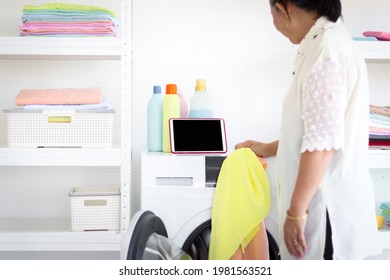 Asian Senior Elderly Old Woman Housewife Doing Laundry At Laundry Room During Looking At Tablet, Grandma Using Tablet In Learning New Things While Doing Housework And Cleaning Clothes At House.