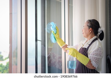 Asian Senior Elderly Woman Housewife Wearing Rubber Gloves And Holding Cleaning Spray And Clothe For Wiping Window, Cleaning Up House, Grandma Doing Housework And Wipeing The Glass.