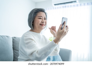 Asian Senior Elderly Woman Smile And Video Call In Living Room At Home. Strong Elderly Older Grandmother Feeling Happy Using Mobile Cell Phone Communicate With Family Enjoy Retirement Life In House.