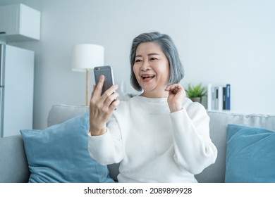 Asian Senior Elderly Woman Smile And Video Call In Living Room At Home. Strong Elderly Older Grandmother Feeling Happy Using Mobile Cell Phone Communicate With Family Enjoy Retirement Life In House.