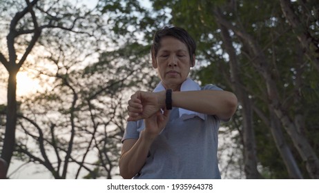 Asian Senior Elderly Woman Touching Smart Watch Activity Tracker At Park Under Tree. Finger Tapping Sport Watch For Calories And Heart Rate Information. Slow Motion Orbit Shot. Health Care Concept.