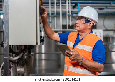 Asian Senior Engineer Wear Safety Vest With Helmet Holding Tablet Checking Electrical Control Cabinet For Machines In Food And Beverage Factory Industry