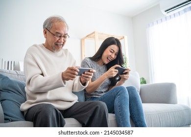 Asian Senior Father And Daughter Play Game On Phone Together In House. Elderly Male And Granddauther Sit On Sofa In Living Room, Feeling Happy To Spend Time Together Playing Game In Smartphone At Home