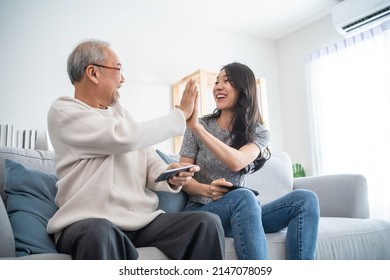 Asian Senior Father And Daughter Play Game On Phone Together In House. Elderly Male And Granddauther Sit On Sofa In Living Room, Feeling Happy To Spend Time Together Playing Game In Smartphone At Home