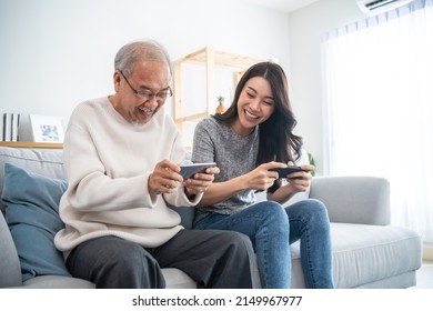 Asian Senior Father And Daughter Play Game On Phone Together In House. Elderly Male And Granddauther Sit On Sofa In Living Room, Feeling Happy To Spend Time Together Playing Game In Smartphone At Home