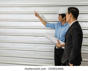 Asian Senior Good-looking Boss In Suit Standing And Working With Junior Employee Wearing White Safety Helmet And Holding Roll Of Paper. Businessmen Teamwork And Good Colleagues Concept.