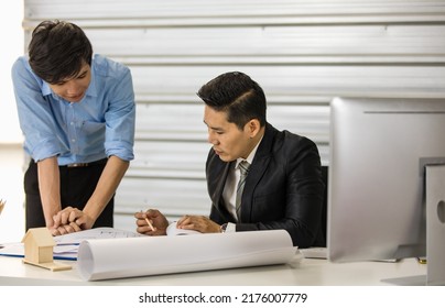 Asian Senior Good-looking Boss In Suit Sitting At Desk And Working With Junior Employee In Office With Roll Of Paper. Businessmen Teamwork And Good Colleagues Concept.