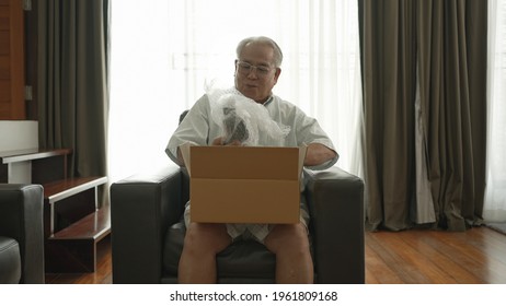 Asian Senior Happy After Retired, Old Man Open Parcel From Shopping Online. Smiling Old Man Open Cardboard Box Sitting On The Sofa In The Living Room Happily With Life Retired.