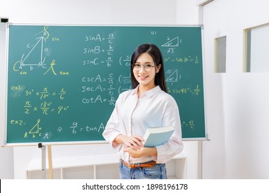 asian senior high school female math teacher hold books and smile at you in front of blackboard at classroom - Powered by Shutterstock