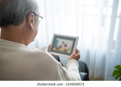 Asian senior male sit alone on wheelchair and looks family photographs. Elderly older mature grandfather feel lonely and sad, missing his relative and wife while stay in nursing home after retirement.