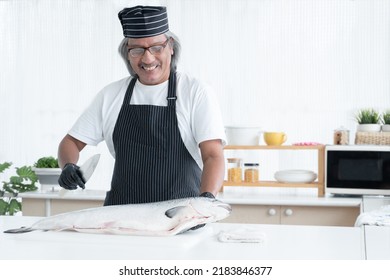 Asian senior man chef is skillfully holding a knife to cut whole fresh salmon in his hand at kitchen. Happy elderly chef prepare good and quality raw ingredients in japanese food - Powered by Shutterstock