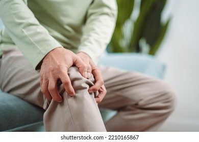 Asian Senior Man Feel Knee Pain Ache. Older Mature Male Hurt At Knee While Sitting On Sofa In Living Room At Home. Bone Problam Health Care And Medical Insurance Concept.